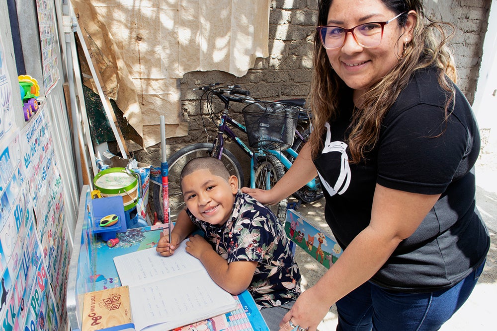 Ramsés fazendo lição de casa com sua mãe, Rosa atrás dele