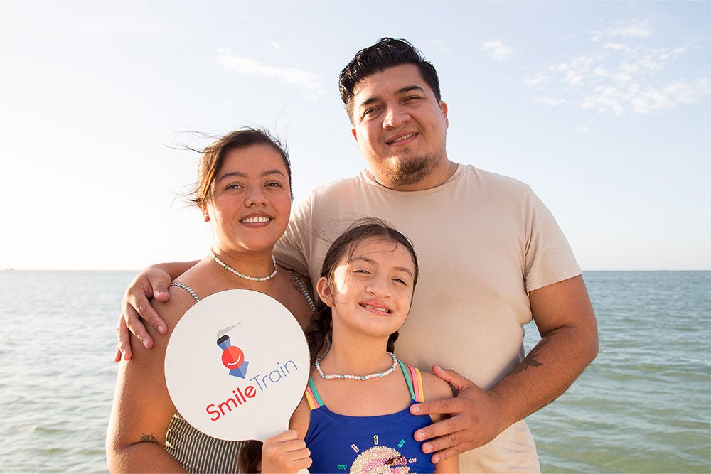 Aquetzaly smiling and holding a Smile Train sign with her parents