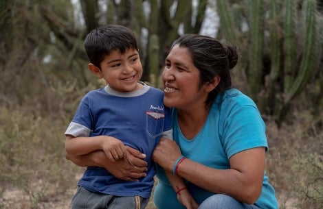 Walter smiles as Valeria kneels and wraps her arms around his affectionately. Behind them are shrubs and cacti
