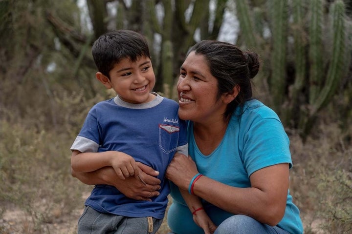 Walter smiles as Valeria kneels and wraps her arms around his affectionately. Behind them are shrubs and cacti
